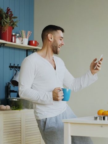 Cheerful happy man dancing and singing in kitchen while browsing social media on his smartphone at home in the morning